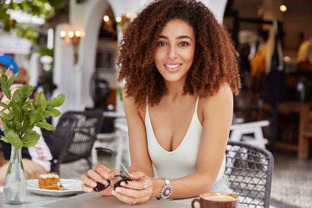 Foto horizontal de uma adorável modelo feminina de pele escura com penteado afro encaracolado, aproveitando o tempo de recreação durante o fim de semana, posa diante do interior aconchegante do café