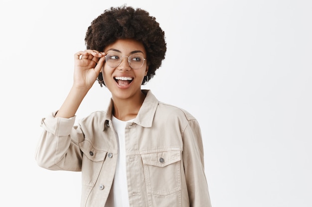 Foto grátis foto horizontal de uma afro-americana lésbica despreocupada e confiante de óculos e camisa bege tocando a borda dos óculos e olhando para a direita, sorrindo amplamente confiante e feliz