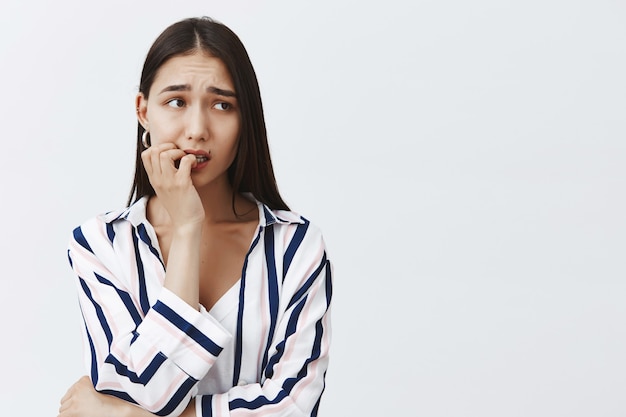 Foto horizontal de uma aluna ansiosa e bonita em uma blusa listrada, roendo a unha nervosamente, franzindo a testa e olhando para o lado, com medo das consequências, em pé sobre uma parede cinza