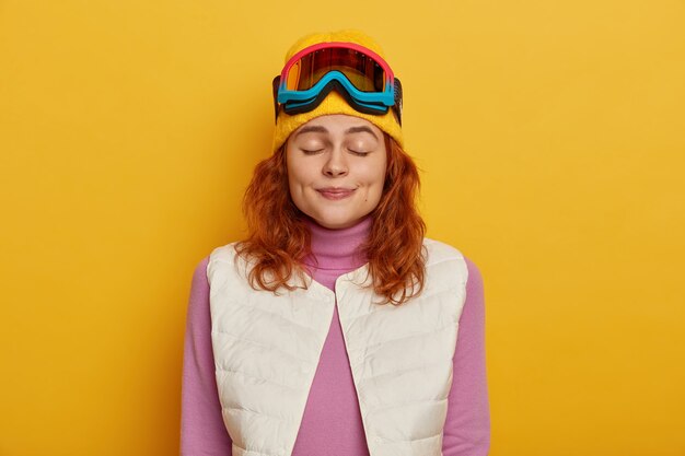 Foto horizontal de uma bela jovem em pé de olhos fechados, vestida com roupas quentes e confortáveis, usando máscara de snowboard para esquiar, isolada sobre fundo amarelo