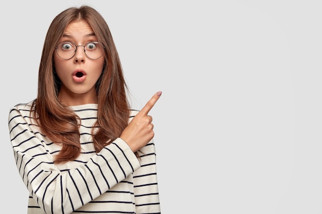 Foto grátis foto horizontal de uma bela jovem mulher branca com expressão de surpresa, aponta para o canto superior direito, vestida com um suéter listrado, usa óculos redondos, isolada sobre a parede branca