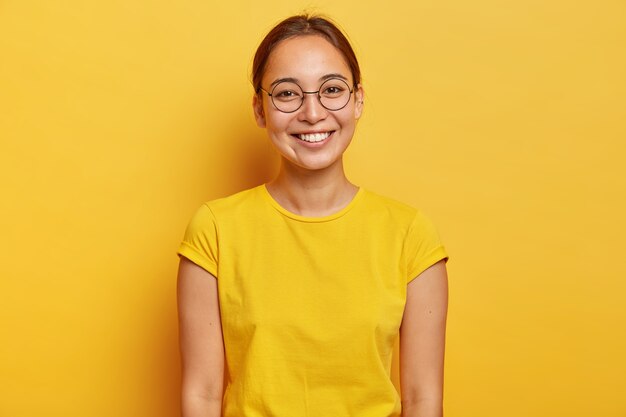 Foto horizontal de uma estudante asiática feliz usando grandes óculos redondos, casual wear amarelo, sorri suavemente, satisfeita após um dia de sucesso na universidade, vestida com uma camiseta amarela de verão