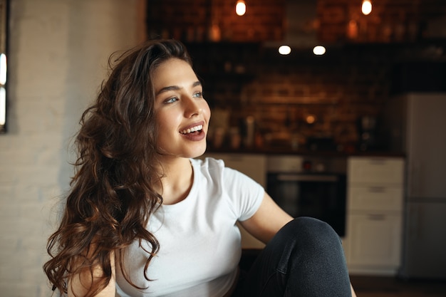 Foto grátis foto horizontal de uma garota bonita com longos cabelos cacheados e olhos azuis sentada confortavelmente na mesa da cozinha, abrindo a boca, cantando, estando de bom humor, se divertindo, parecendo com um sorriso feliz e sonhador