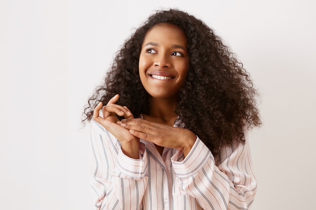 Foto horizontal de uma jovem afro-americana bonita em uma camisola elegante, olhando para cima com uma expressão facial pensativa e animada, mordendo o lábio e esfregando as mãos, tendo uma ideia ou um plano brilhante, sonhando