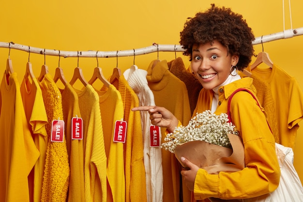 Foto horizontal de uma jovem afro-americana encantada aponta para roupas elegantes à venda penduradas nos trilhos, carrega uma bolsa e um lindo buquê, tem um sorriso cheio de dentes, isolado sobre um fundo amarelo