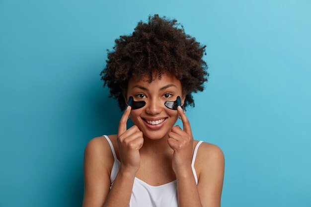 Foto horizontal de uma jovem afro-americana se preocupando com a pele, aponta para manchas hidratantes antienvelhecimento de hidrogel sob os olhos, gosta de tratamento de beleza, tem um sorriso largo, isolado na parede azul