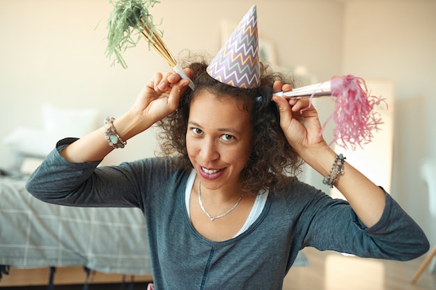 Foto horizontal de uma jovem feliz mestiça com chapéu cônico na cabeça, se divertindo e brincando
