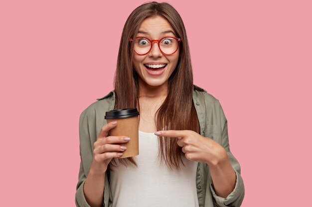 Foto horizontal de uma jovem morena alegre aponta para um café para viagem, tem uma expressão alegre, anuncia uma bebida aromática,