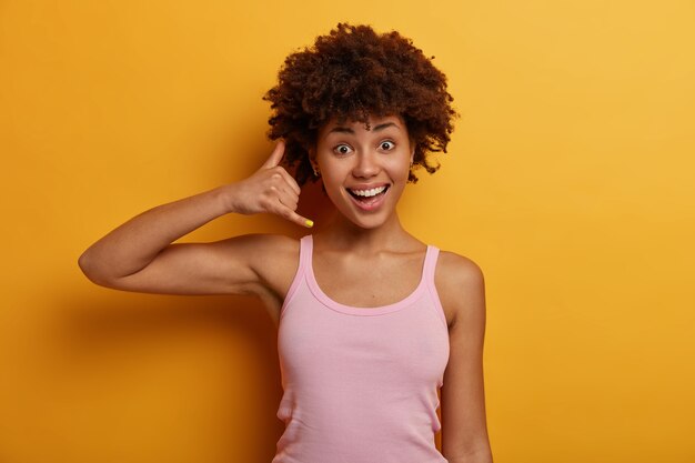 Foto horizontal de uma jovem simpática e alegre faz gestos de telefone, pede para ligar para ela, sorri positivamente, mantém contato com os amigos, tem cabelo encaracolado, posa dentro de casa contra uma parede amarela