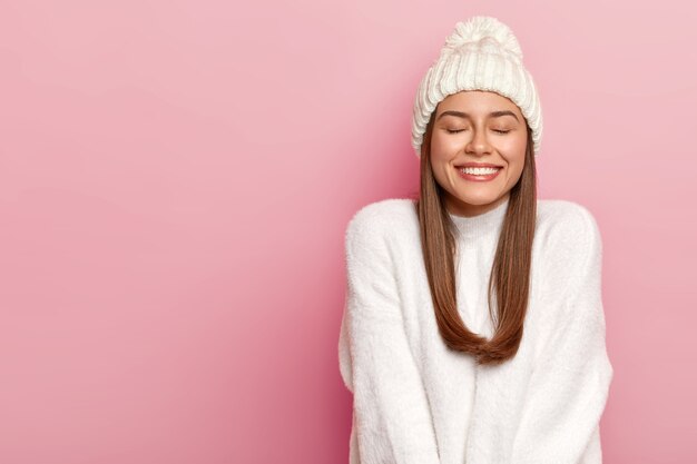 Foto horizontal de uma linda jovem com cabelo escuro, mantém os olhos fechados, sorri agradavelmente, mostra dentes brancos perfeitos, desfruta de conforto com um suéter recém-comprado, chapéu quente