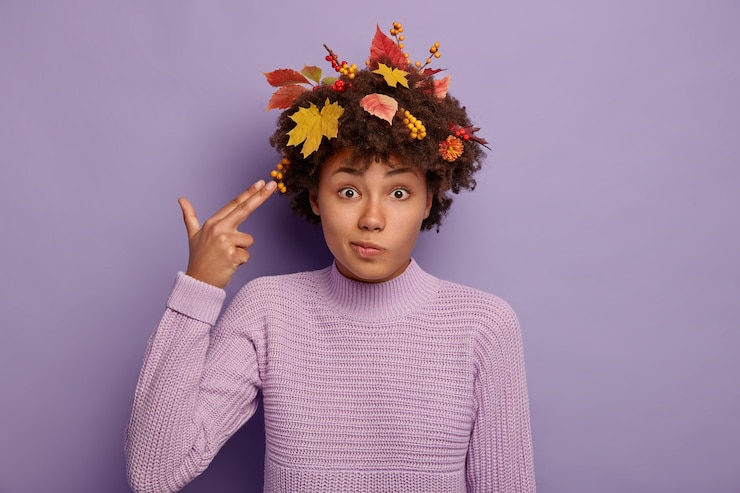 Foto grátis foto horizontal de uma linda mulher afro-americana perplexa atira na têmpora com os dedos, mostra um gesto suicida, vestida com um suéter quente, enquanto folhas amarelas no cabelo