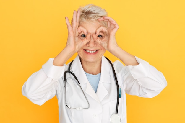 Foto horizontal de uma mulher atraente, alegre, de meia-idade, médica, vestindo jaleco branco e estetoscópio ao redor do pescoço, se divertindo, mostrando uma atitude positiva, fazendo binóculos com os dedos, sorrindo