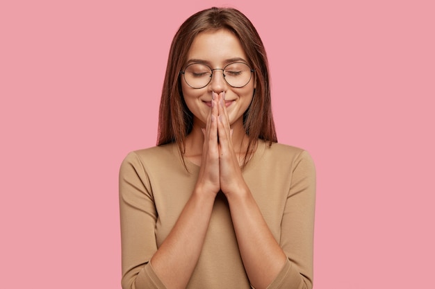 Foto grátis foto horizontal de uma mulher atraente e satisfeita orando por um bem-estar, mantendo as palmas das mãos pressionadas uma contra a outra