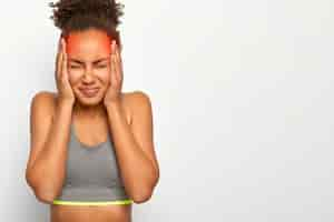 Foto grátis foto horizontal de uma mulher de cabelo encaracolado insatisfeita tocando ambas as têmporas, sofre de enxaqueca, usa sutiã esportivo, fecha os olhos devido a sentimentos ruins, isolado na parede branca