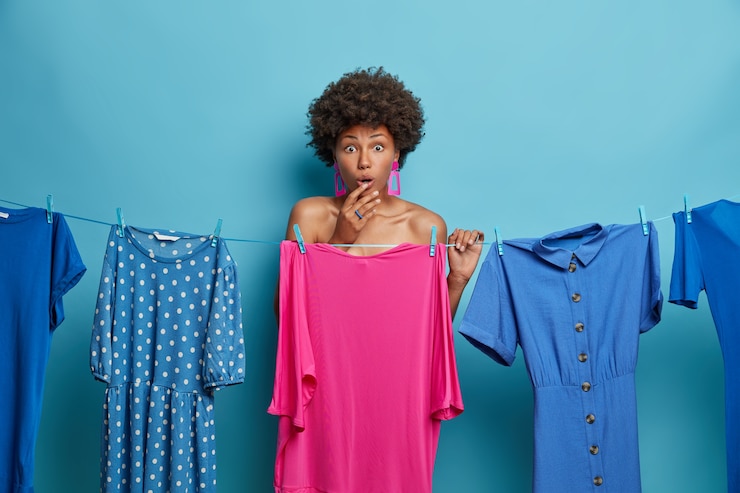 Foto grátis foto horizontal de uma mulher de pele escura surpresa enfrenta problemas com o que vestir, fica atrás do vestido rosa, tem uma expressão estupefata, posa perto da corda com vestidos lavados pendurados. hora de se vestir.