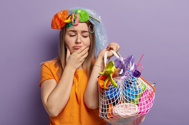 Foto horizontal de uma mulher europeia insatisfeita com expressão facial triste, segura um saco de resíduos de polietileno, segura o queixo, deprimida com o meio ambiente destruído e sério problema de poluição