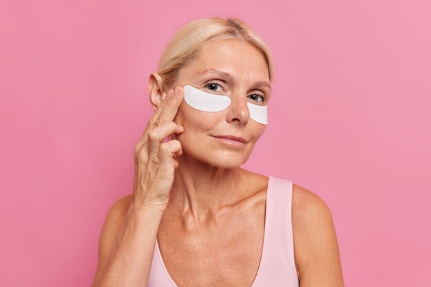 Foto horizontal de uma mulher loira de quarenta anos aplica manchas de beleza sob os olhos e passa por procedimentos anti-envelhecimento olha atentamente para a câmera isolada sobre a parede rosa