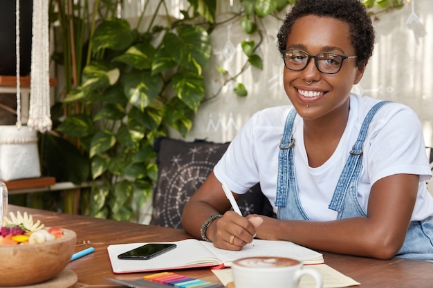 Foto horizontal de uma mulher negra alegre usando óculos, escrevendo listas para fazer em um caderno ou diário pessoal