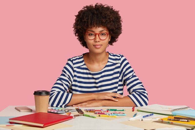 Foto horizontal de uma mulher negra atraente com cabelo crespo, com expressão séria, sentada na mesa branca, fazendo ilustrações em um caderno espiral, vestida com um macacão casual listrado, óculos ópticos