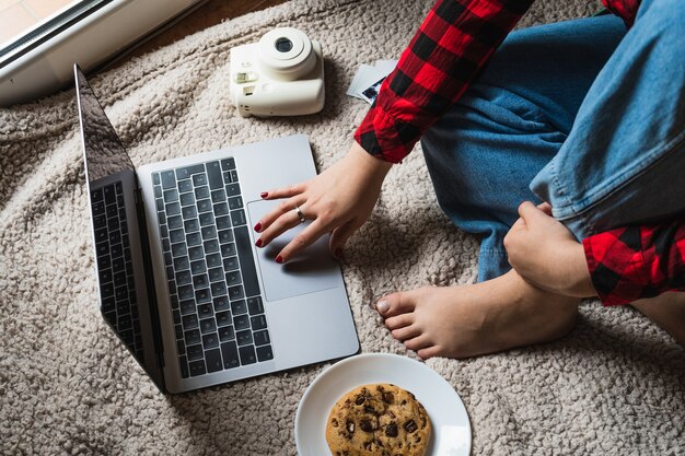 Foto horizontal de uma mulher sentada em um cobertor com seu laptop e um biscoito