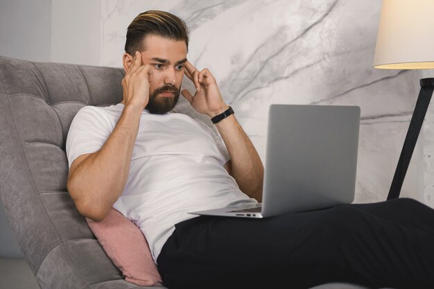 Foto horizontal interna de um jovem empresário com a barba por fazer deprimido e frustrado usando um computador portátil para trabalho remoto, sentindo-se estressado por causa de problemas, massageando as têmporas, tendo fortes dores de cabeça