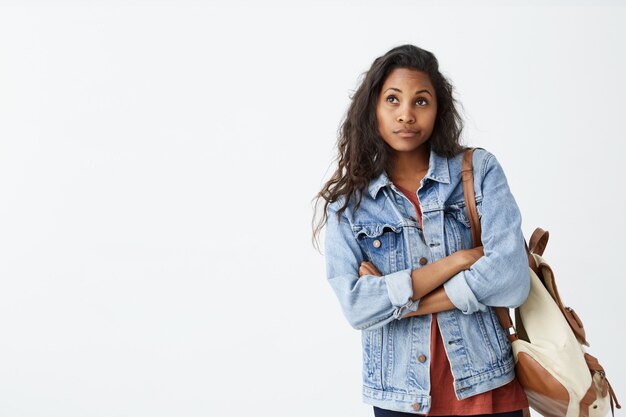 Foto interior da linda garota de pele escura, olhando para longe, tendo expressão no rosto duvidosa e indecisa, perseguindo os lábios como se ela não estivesse satisfeita. Fêmea jovem confusa na jaqueta jeans com mochila