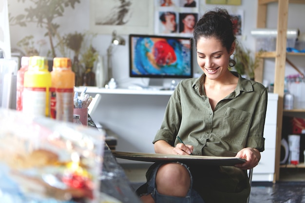 Foto interior de feliz jovem artista feminina caucasiana feliz sorrindo amplamente enquanto trabalhava na pintura ou desenhos na oficina; material de pintura