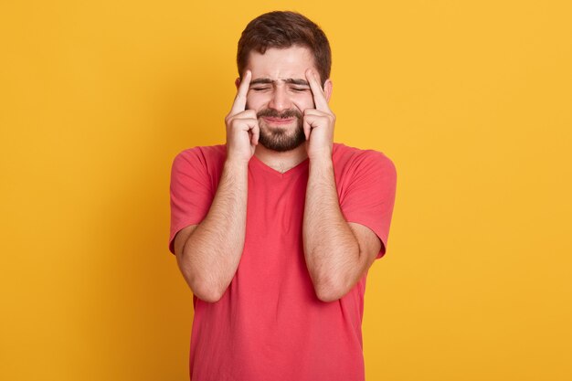 Foto interior de rosto descontente masculino, com dor de cabeça, sensação de cansaço, precisa de descanso, fecha os olhos com dor, mantém os dedos nas têmporas