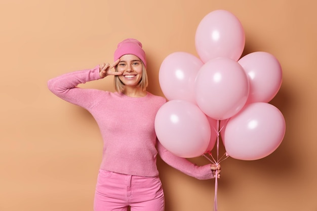 Foto interior de uma garota alegre e bonita faz um gesto de paz sobre o olho segura um monte de balões de hélio inflados vem na universidade saindo da festa usa um jumper de chapéu rosa e jeans parece alegre