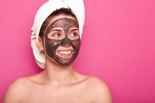 Foto interior de uma jovem mulher atraente com uma toalha branca na cabeça, tem o corpo nu, sorrindo isolado em rosa no estúdio, olha de lado, com uma máscara de chocolate no rosto. Conceito de skincare.