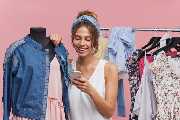 Foto interior do adorável comprador feminino, gastando seu tempo livre na boutique, perto de manequim com roupas, lendo notícias on-line enquanto estiver usando a conexão gratuita à Internet. Assistente de loja que vende roupas