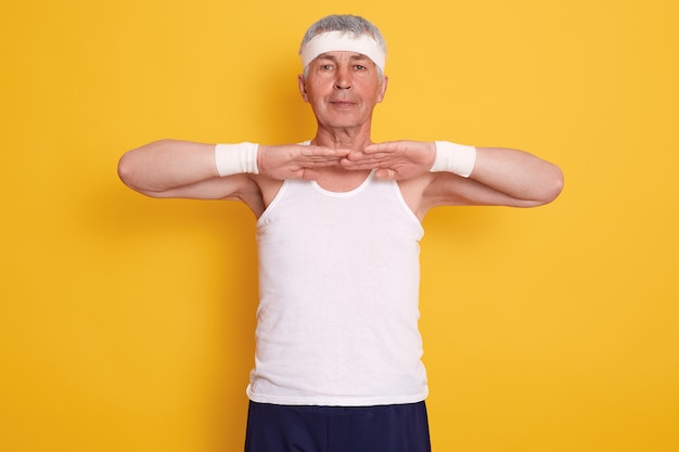 Foto interior do homem desportivo sênior, vestindo camiseta sem mangas e bandana, mantendo as mãos na frente do peito, fazendo exercícios físicos