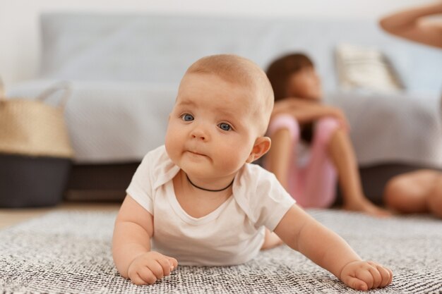 Foto interna de bebê fofo vestindo roupas brancas, deitado no chão, no tapete, em sua barriga, estudando o mundo ao redor por conta própria, olhando para a câmera com uma expressão curiosa.