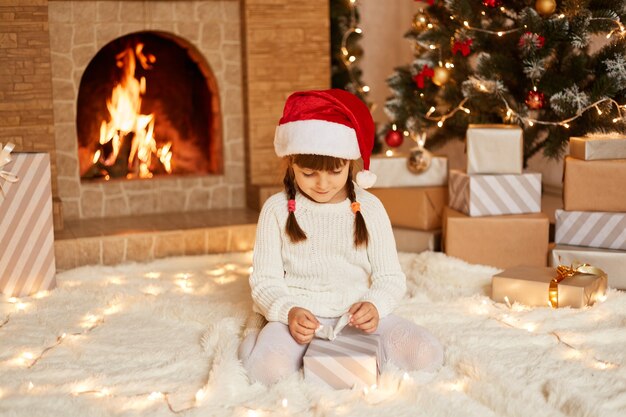 Foto interna de encantadora criança do sexo feminino com suéter branco e chapéu de Papai Noel, abrindo a caixa de presente do Papai Noel, posando na sala festiva com lareira e árvore de Natal.