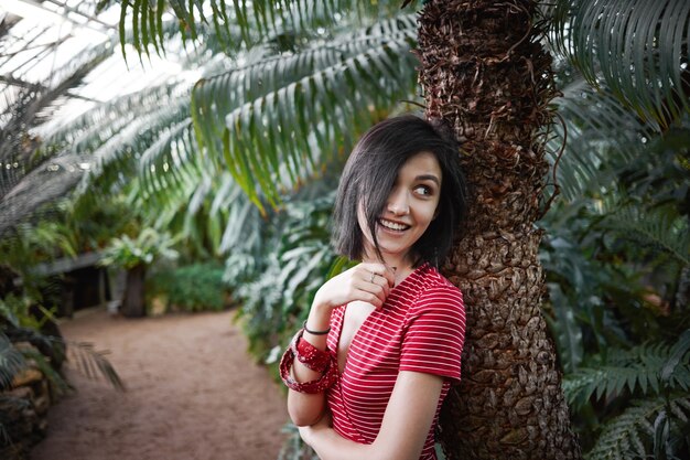 Foto interna de encantadora jovem fêmea com sorriso fofo e cabelo curto escuro, encostado na grande palmeira, olhando por cima do ombro e sorrindo divertidamente. Conceito de pessoas, natureza, estilo e moda