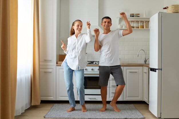 Foto interna de marido e mulher felizes e positivos dançando, se divertindo juntos na cozinha, celebrando a mudança, estando de bom humor e expressando felicidade.