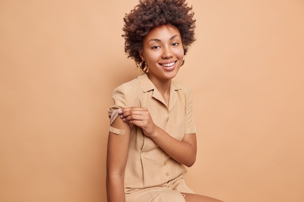 Foto interna de mulher positiva com cabelo encaracolado mostra braço vacinado com gesso recebendo segunda dose da vacina feliz por se sentir protegido levanta a manga do vestido isolada sobre a parede bege