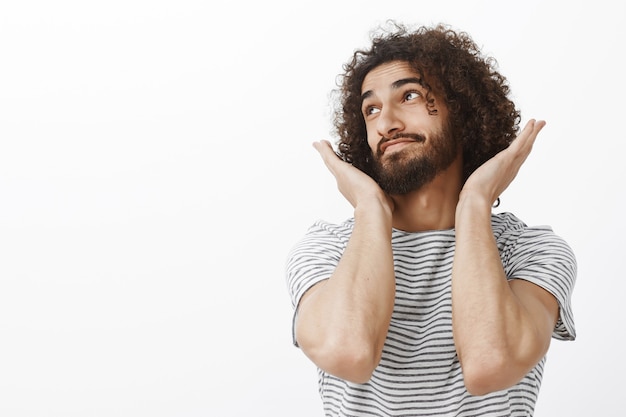 Foto grátis foto interna de um cara barbudo bonito e despreocupado com penteado afro, olhando para longe e sorrindo com uma expressão sonhadora, levantando as palmas das mãos e segurando-o perto do queixo