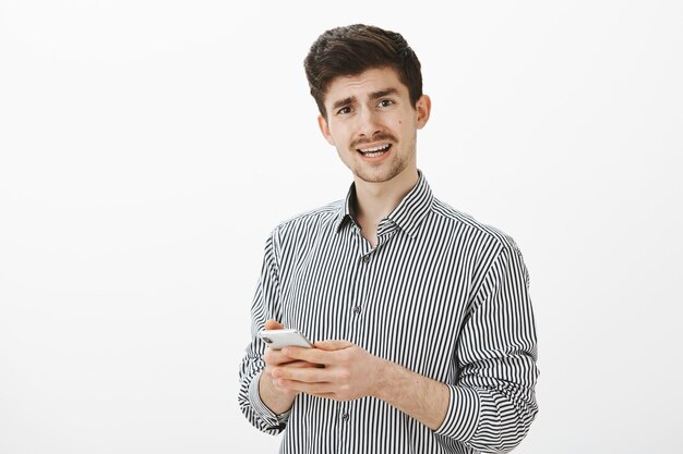 Foto interna de um cara maduro descontente e frustrado com bigode em uma camisa listrada casual, parecendo questionado ao fazer uma pergunta e segurando um smartphone, recebendo uma mensagem confusa