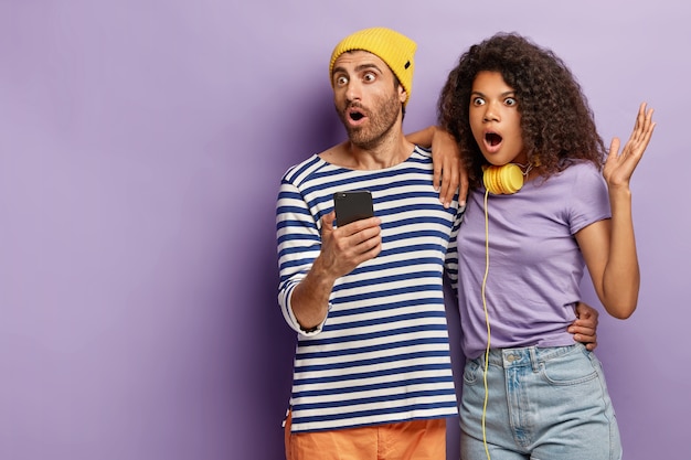 Foto interna de um casal assustado e estupefato checando notícias no celular, olhando para longe, olhando para a cena terrível