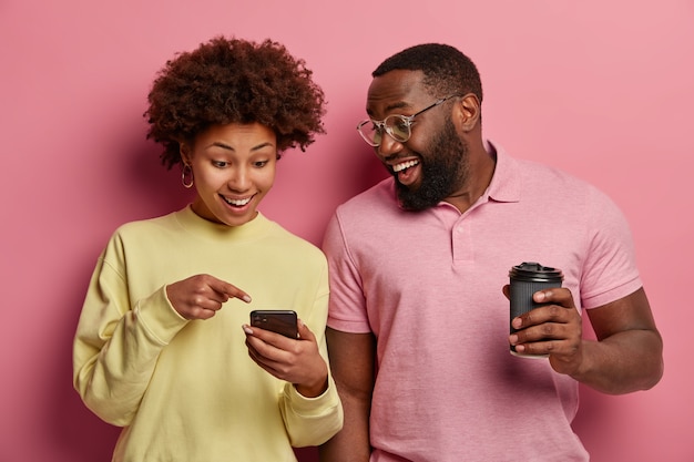 Foto interna de um casal feliz de pele escura tomando café para viagem, assistindo ou lendo informações on-line em um gadget moderno e vestindo roupas em tons pastéis