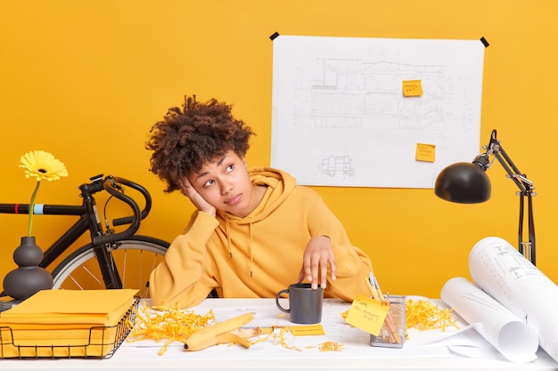 Foto grátis foto interna de um estudante afro-americano atencioso se preparando para os exames sonhos com feriados e poses de descanso na dekstop com papéis adesivos esboços vestidos com moletom amarelo casual tem cursos de design