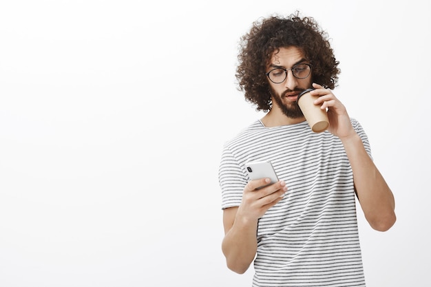 Foto interna de um estudante masculino bonito interessado em óculos da moda e camiseta listrada, enviando mensagens pelo novo smartphone branco, bebendo café na xícara