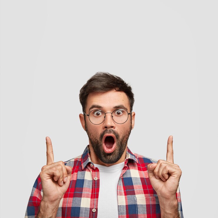 Foto grátis foto interna de um homem barbudo perplexo aponta para cima, sendo surpreendido enquanto posa contra uma parede branca.