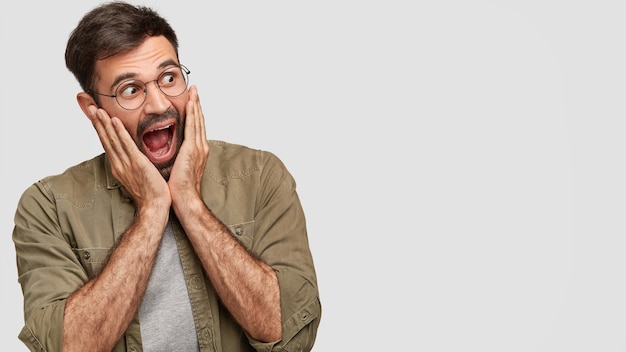 Foto interna de um homem caucasiano estupefato e surpreso com a barba por fazer, mantém o queixo caído, toca as bochechas com as palmas das mãos, reage a algo