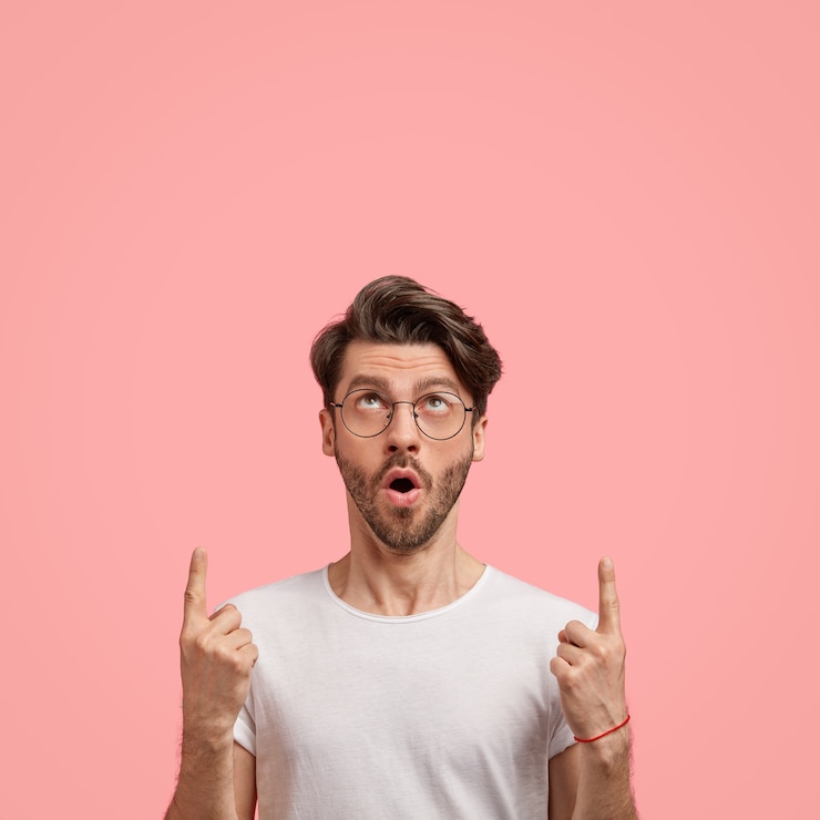 Foto grátis foto interna de um homem com a barba por fazer emotivo com penteado moderno, cerdas escuras, abre a boca de maravilha, aponta com os dois dedos indicadores, mostra espaço livre para seu conteúdo promocional, vestido casualmente
