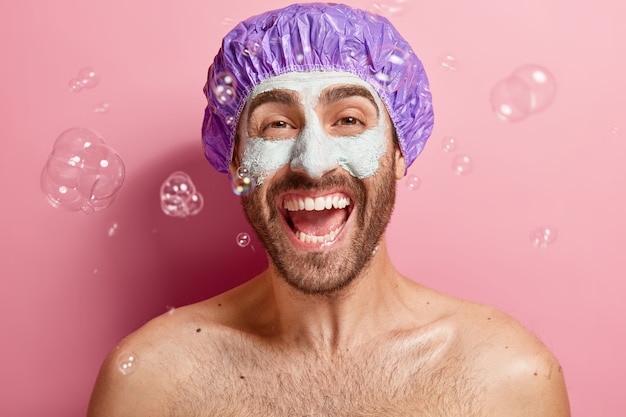 Foto interna de um homem emocionado e satisfeito com máscara de argila, gosta de tomar banho e fazer tratamento facial, usa touca de banho, bolhas de sabão voando por aí, lava o corpo