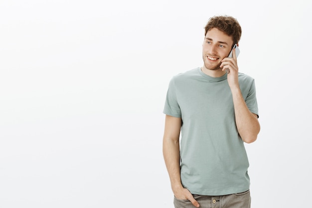 Foto interna de um homem europeu charmoso e relaxado com cabelo loiro, segurando o smartphone perto da orelha e olhando para a esquerda com um largo sorriso, conversando casualmente com a irmã