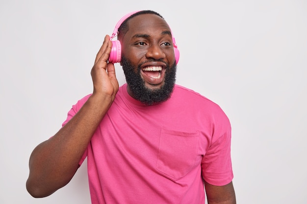 Foto interna de um homem negro feliz com clima otimista ouve música por meio de fones de ouvido sem fio curtindo a playlist favorita parece distante com expressão alegre vestida casualmente isolada na parede branca