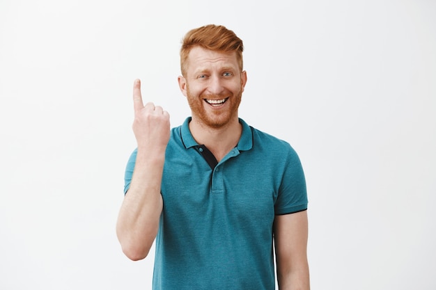 Foto interna de um homem ruivo bonito triunfando e satisfeito com a cerda mostrando o dedo indicador no gesto número um ou primeiro, sorrindo e rindo alegremente sobre a parede cinza